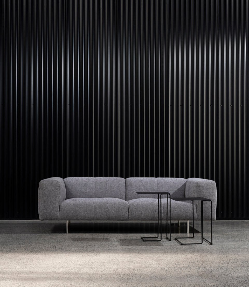 Milano plush sofa in grey upholstery, paired with two minimalist black Revello side tables on a polished concrete floor