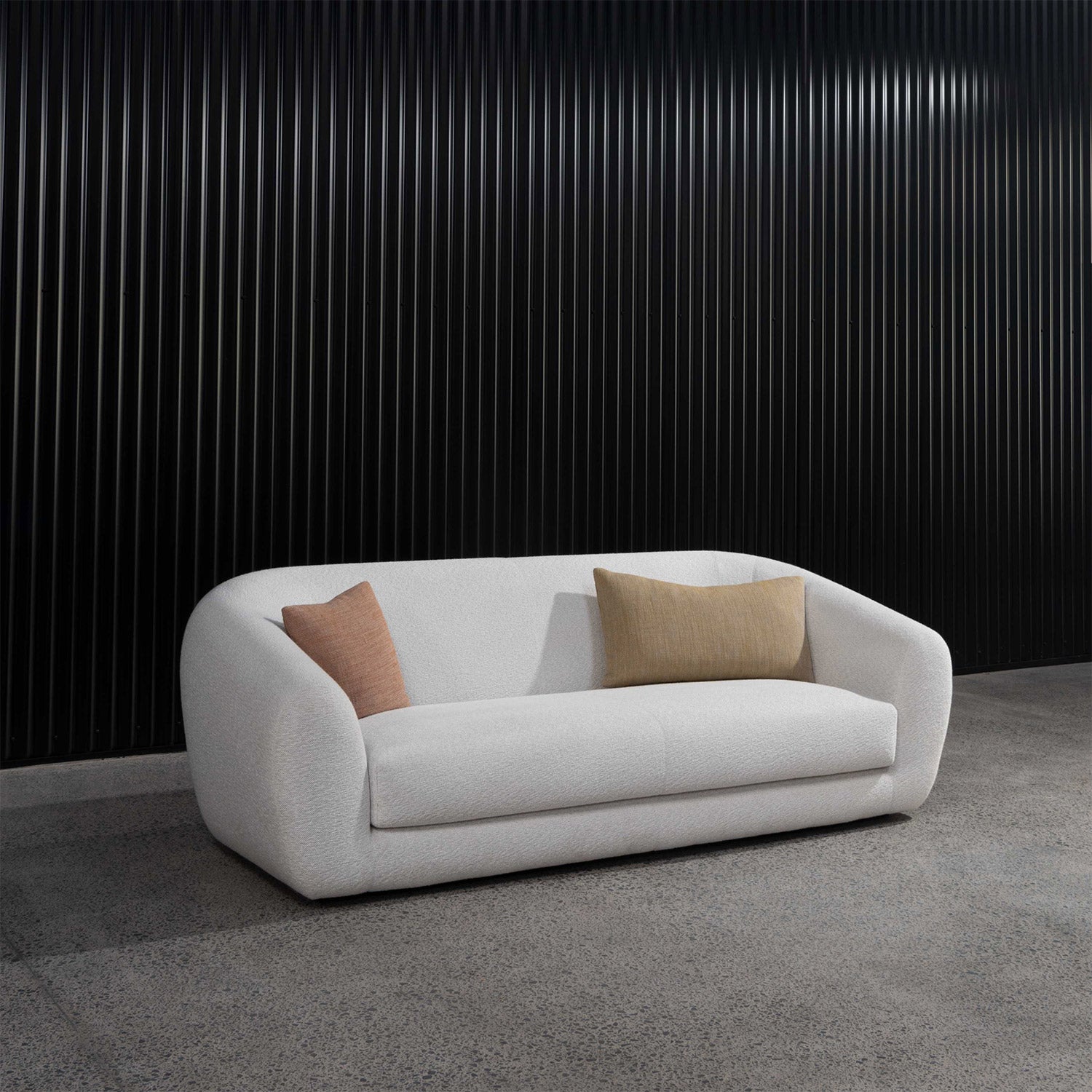 Pisa Plush 2-seater sofa in cream colour with two cushions on showroom floor. Black wall background.
